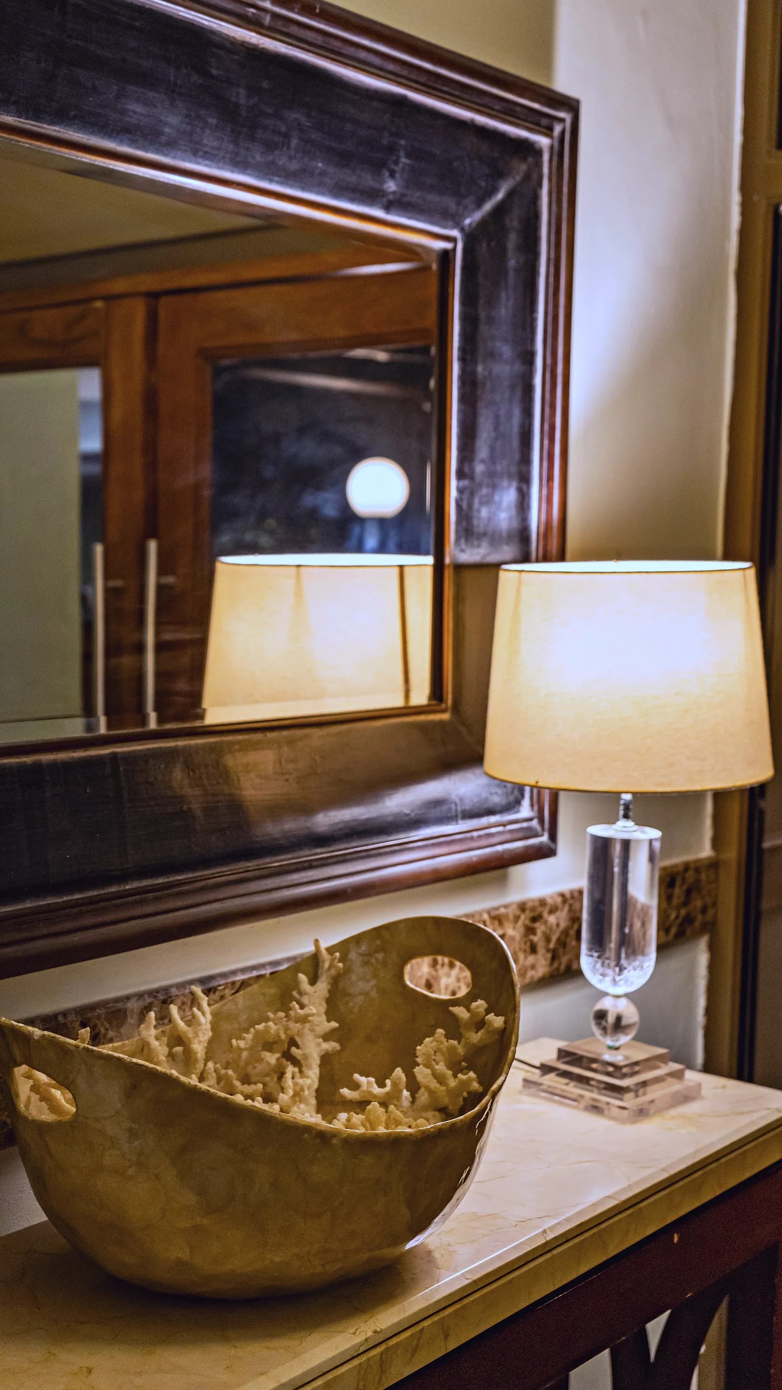 An antique mirror, glowing table lamp and coral-filled bowl on a console table at The Alcove